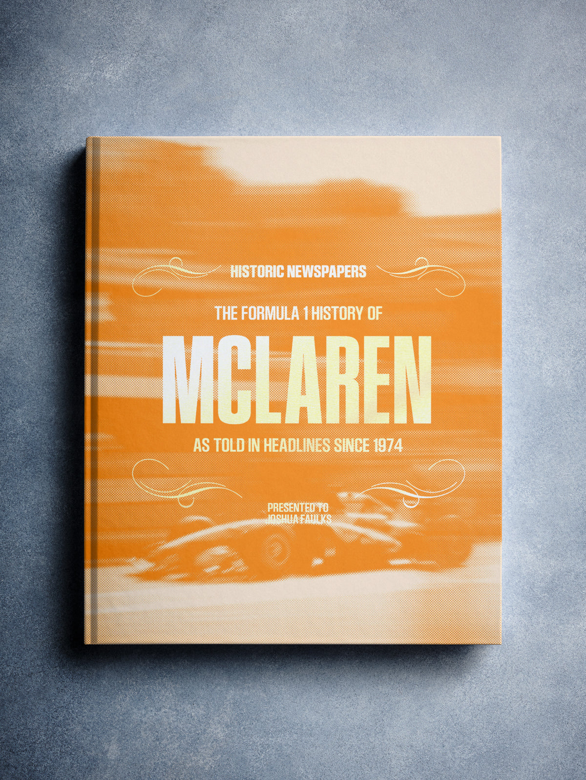 Close-up of "The Formula 1 History of McLaren" POD book with orange foil cover, showcasing detailed cover design and branding.